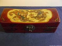 Front angled photo of the red lacquer rectangular box with domed lid showing colorful painted oriental dragons and floral accents on a cream inset panel.