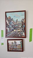 Two paintings on wall: upper large city street scene with tram, lower smaller architectural cityscape.