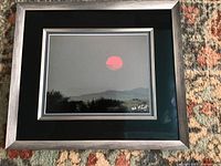 Framed artwork showing red sun over a dark landscape with hills in the background. The frame is silver with a black or dark green mat.