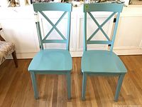Front view of two blue wooden chairs with crossed back design, on a hardwood floor.
