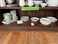 Wide shot of the shelf showing multiple pieces of floral patterned dinnerware including stackable soup bowls, small and large plates, and an oval serving bowl under a cabinet shelf
