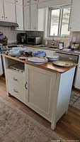 Full view of kitchen island cabinet with white body and wood top, drawers and door visible, some plates on top.