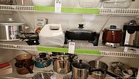 Kitchen shelf with various small kitchen appliances including black Cuisinart waffle maker, brown ceramic West Bend electric bean pot, black rectangular Elite Gourmet electric cooker, white Hamilton Beach hand mixer, stainless steel pots and measuring cup