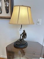 Full view of the lamp showing hunter and dog figure with beige lampshade, on black oval base. Lamp is on.