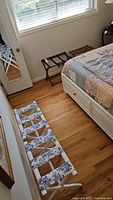 Four luggage stands in a bedroom near bed and window. White wooden luggage stand with blue and white floral fabric straps in foreground and smaller wooden luggage stand with decorative floral fabric making a set against the wall.