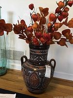 Full view of Marajoara art ceramic vase with faux floral arrangement on wooden floor near wall vent