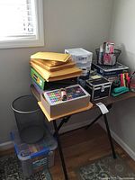 Wide photo of office supplies on black folding table showing padded envelopes, markers, black stapler, storage drawers, tape dispenser, books, and mesh trash can next to table.