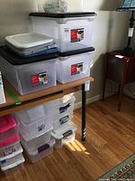 Photo showing multiple translucent plastic storage boxes stacked on a wooden table and floor, mostly with black lids, some with labels visible for Office Depot split-lid file box.
