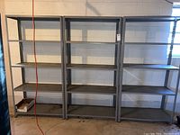 Three metal utility shelf units placed side by side against a basement wall, each with five metal shelves showing signs of rust and dirt.