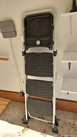 Full view of the folded three-tiered step ladder leaning against a wall, showing aluminum frame and black plastic steps and tray.