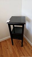 Side view of the dark brown side table showing the drawer and open shelf, standing on a wood floor near a white wall.