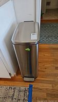 Front angled view of rectangular stainless steel trash can with foot pedal on hardwood floor.