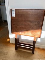 Three wooden TV trays stacked upright against a wall, showing the complete surface of one tray with scratches and light paint marks.