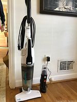 Full view of Simplicity Spiffy upright vacuum and Dirt Devil handheld vacuum side by side in a room with wood flooring and white wall and power outlet