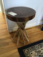 Full view of the round wooden table showing the thick wooden top and natural branch legs crossed as the base, standing on hardwood floor near a wall.