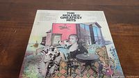 Photo of the front cover of The Hollies Greatest Hits vinyl LP showing detailed illustrated album art with characters and objects.