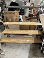 Front view of 3-shelf wooden display with metal frame and caster wheels, showing shelf surfaces and metal handlebar