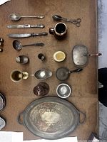 Top view of assorted antique metal dishes and utensils laid out on table surface, includes large tray, various spoons, knives, pot, small pan, scissors, cups, and kettle.