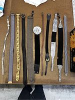 Full view of 11 assorted belts laid side by side on a table showing variety of styles, textures and designs from braided leather to patterned fabric.