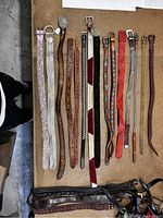 Top down image showing arranged assortment of designer belts including white snakeskin, brown leather, studded black leather, multicolor fabric, red and beige belts.
