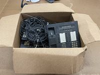 Box filled with assorted black land line telephones and wires packed inside a cardboard box.