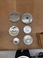Photo showing seven assorted pot lids arranged on floor showcasing different sizes, handles, and materials.