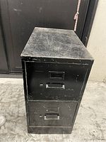 Front side view of black metal filing cabinet with two drawers showing surface scratches and scuffs.