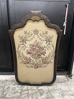 Front view of decorative fireplace screen showing floral needlepoint tapestry inset and scalloped wooden frame with carved shell motif.