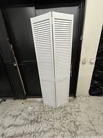 Full view of the white folding privacy screen standing upright against a dark background, showing its three connected hinged panels and slatted design.