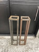 Two tall rectangular metal frame stands placed side by side on a rough floor, showing their full length and rusted surfaces.