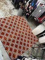 Orange and white geometric patterned rug displayed on floor near clothing racks and umbrella stand.