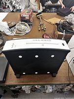 Photo showing the top panel and underside of a white and black paper shredder placed on a brown surface in a cluttered environment with other items around.