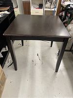 Front view of the tall square wooden table showing its dark surface, tapered legs, and worn condition.