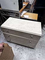 White three-drawer cabinet with marks on top surface, pictured in a cluttered office or storage environment.