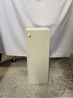 Photo of the front view of the small white cupboard with closed door and round knob visible.