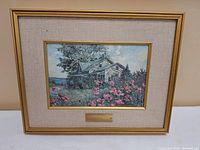 Framed painting showing a house with pink flowers, set in a gold frame with beige mat