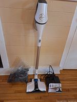 Full view of the Hoover Reach React cordless vacuum standing upright, along with a charging base, user manual, and bag of accessories on the floor.