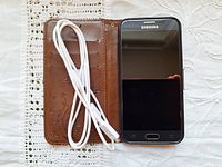 Samsung Galaxy J3 Prime phone with screen facing up, beside a brown folio-style phone case with floral embossing and a white charging cable coiled loosely.