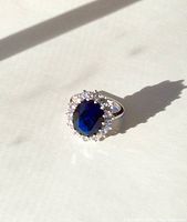 Top angled view of sterling silver Princess Diana style ring showing large blue oval cubic zirconia center stone and surrounding clear cubic zirconia stones in halo setting, casting shadow on white surface.