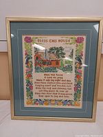 Full view of framed embroidery showing house, birds, flowers and prayer text inside a matted frame.