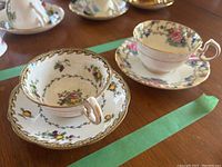 Two Aynsley tea cups and saucers displayed on a table. One set has yellow lemons and purple flowers, the other set has pink and blue floral patterns with gold trim.