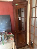 Front view of tall wood veneer cabinet with inset glass door and lower wood door, showing ornate brass hardware and crown molding top.