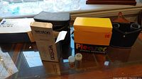 Weston WX-7 camera original box, Pleaser Kodak instant camera box, film canisters, and black leather case for binoculars on table next to window