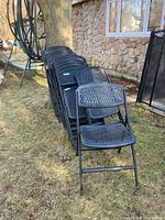 Nine black folding chairs stacked outdoors against a tree and stone house wall