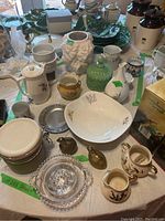 Wide view of assorted ceramic, glass, and metal serving ware and vases arranged on a table including pitchers, bowls, and decorative items.