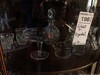 Glass and crystal assortment displayed on wooden cabinet shelf under lighting showing details of cuts and shapes.