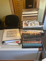 Stack of assorted hardcover books on white surface including home improvement binder and various novels, seen in office setting.