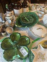 Green ceramic leaf-shaped dishes stacked, Noritake covered casserole dish visible, small cups and bowls arranged on table, white ironstone canisters with dark brown lids.