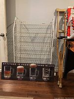 White wire rack shelf with four tiers made of metal wire, slightly worn, shown in front of a wall and next to wooden crutches and a beer sign on the floor.