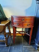 Front view of oval wooden side table showing three drawers with ring-pull handles and circular shelf between legs.